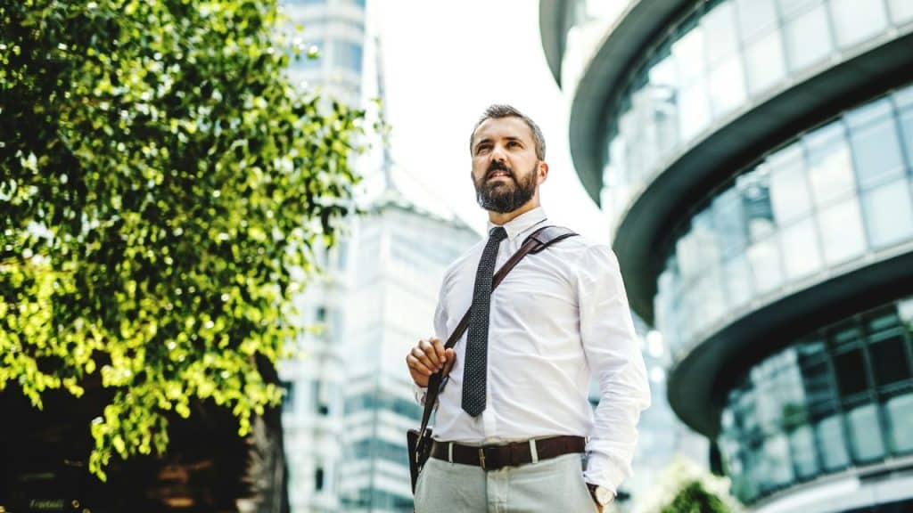 A smiling businessman with a beard and tie stands outdoors against a modern building.