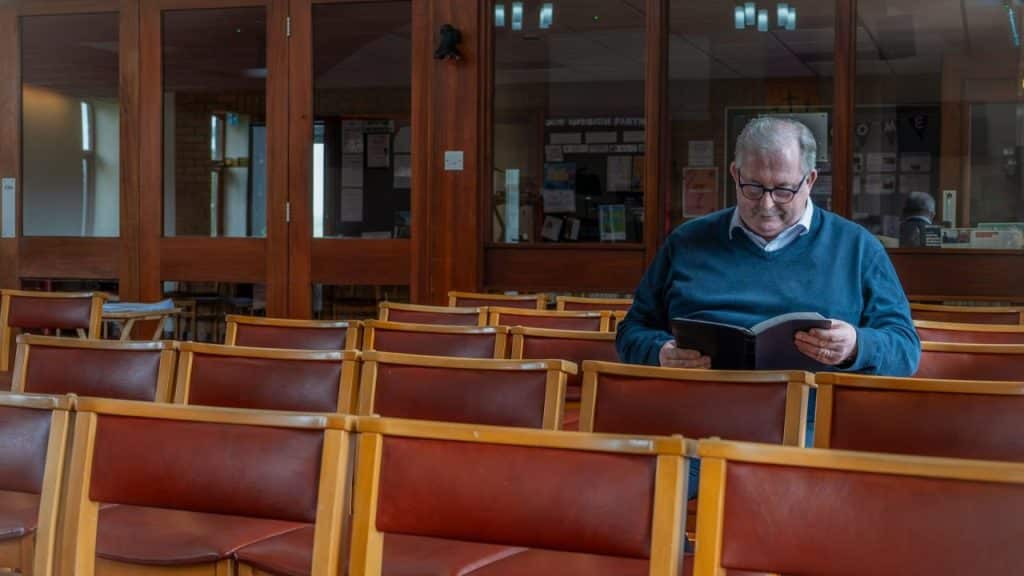 Older man in a blue sweater sitting alone in a row of red and wood chairs, reading a book.