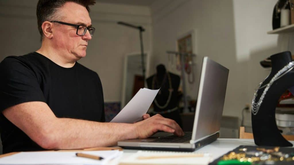 Man in glasses and a black shirt working on a laptop with papers and jewelry nearby.