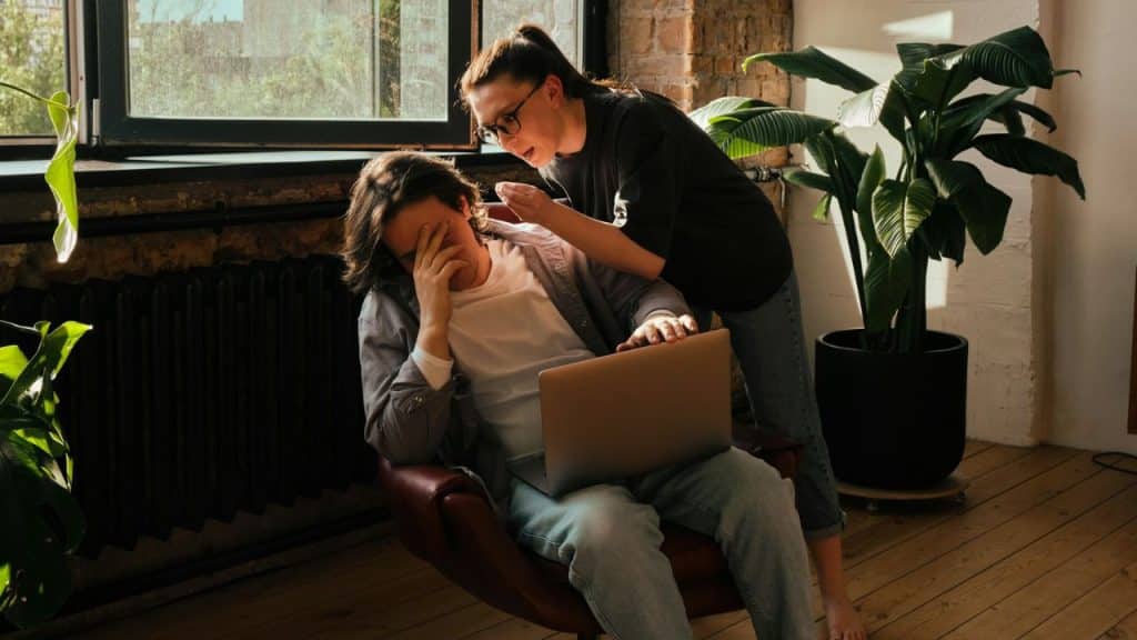 A man sits with a laptop, covering his face while a woman talks to him.