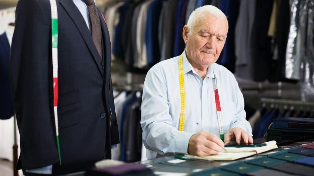 Elderly tailor with a measuring tape writing in a notebook next to a mannequin.