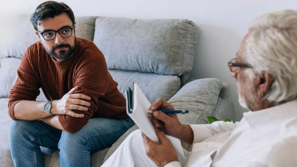 Younger man with a beard sits on a couch across from an older man writing on a clipboard.