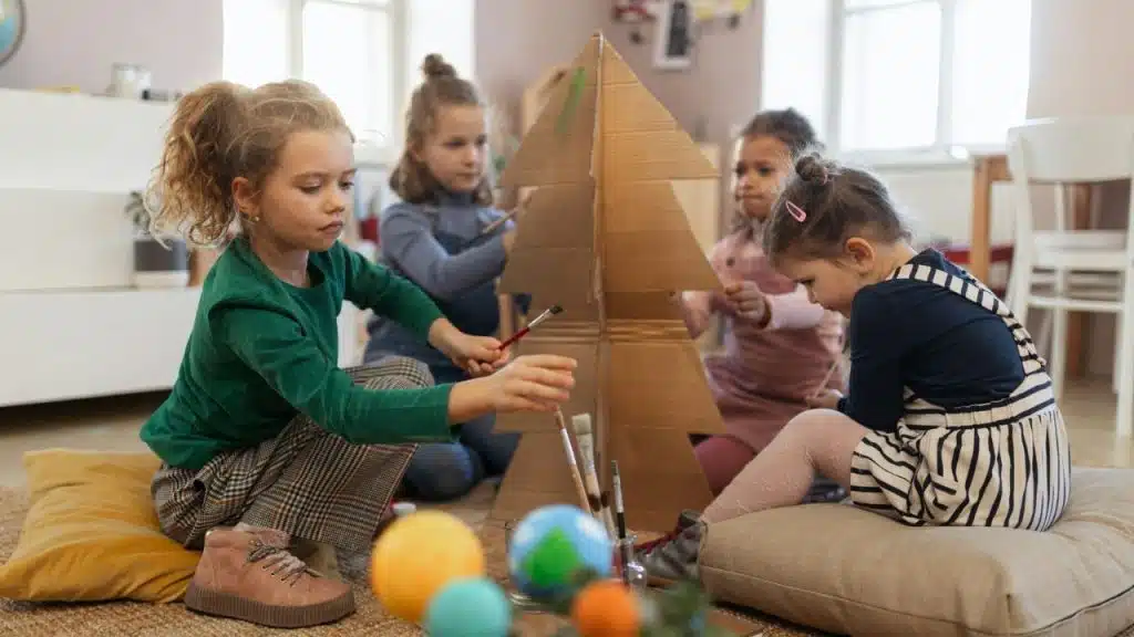 Four young girls painting a large cardboard Christmas tree in a sunlit room.