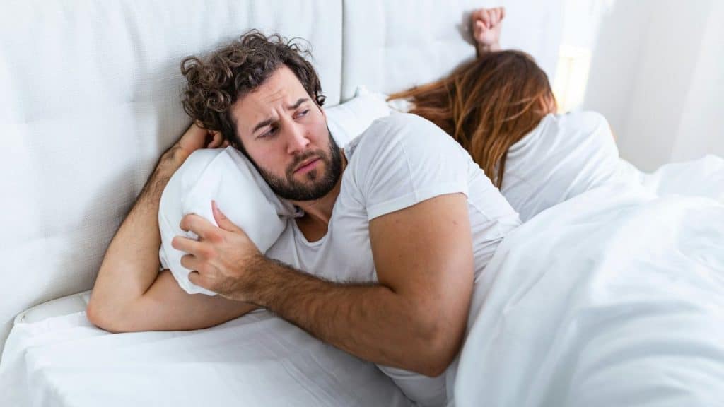 A distraught man with a beard lies in bed, turning away from the woman next to him.