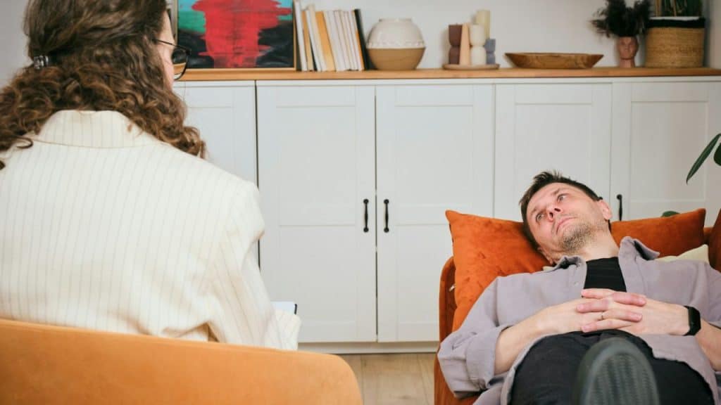 A man lies back on an orange couch, looking up, while a woman with curly hair sits in a chair facing him.
