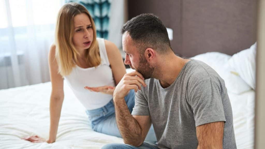 A blonde woman with a frustrated expression gestures toward a man sitting on a bed who appears deep in thought.