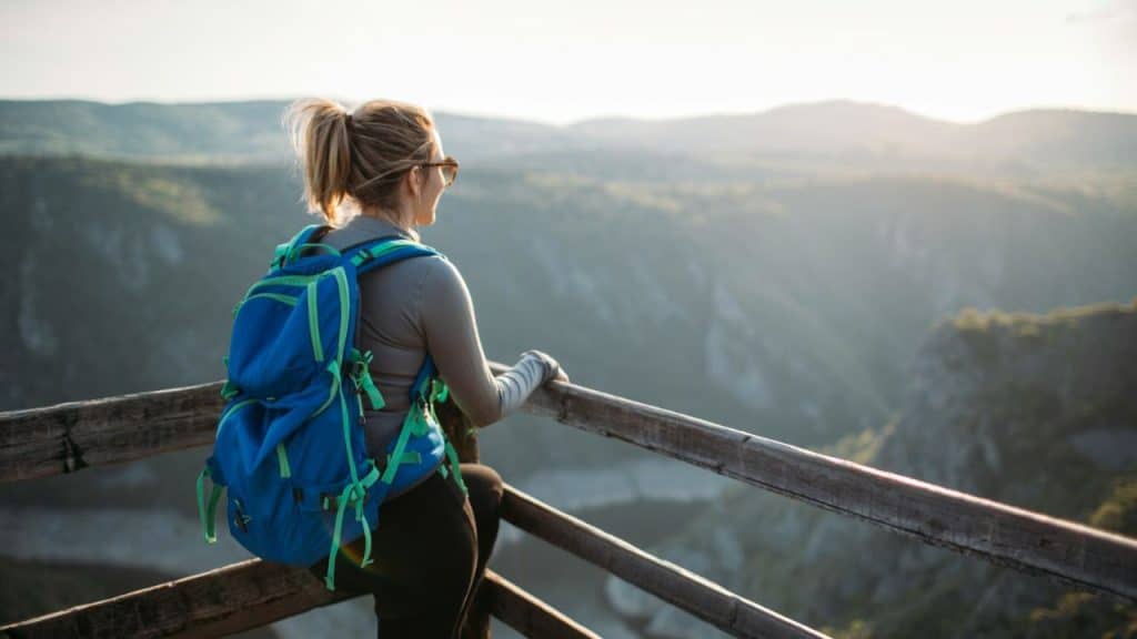 A woman with a blue backpack stands on a wooden platform overlooking a valley at sunset.