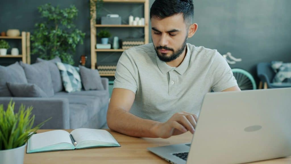 A focused man with a beard sits at a wooden desk working on a laptop in a modern living room.