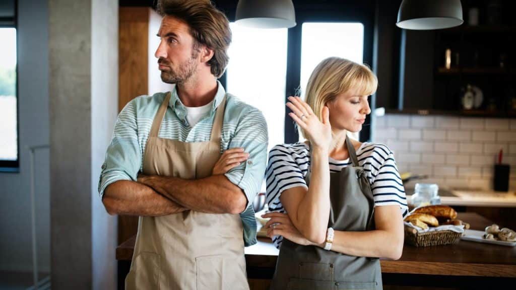 Man and woman in aprons standing back-to-back in a kitchen, looking upset.
