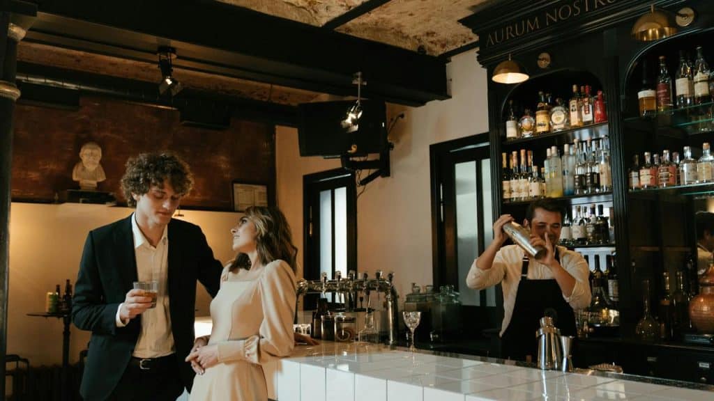 A couple standing at a bar while a bartender mixes a drink.