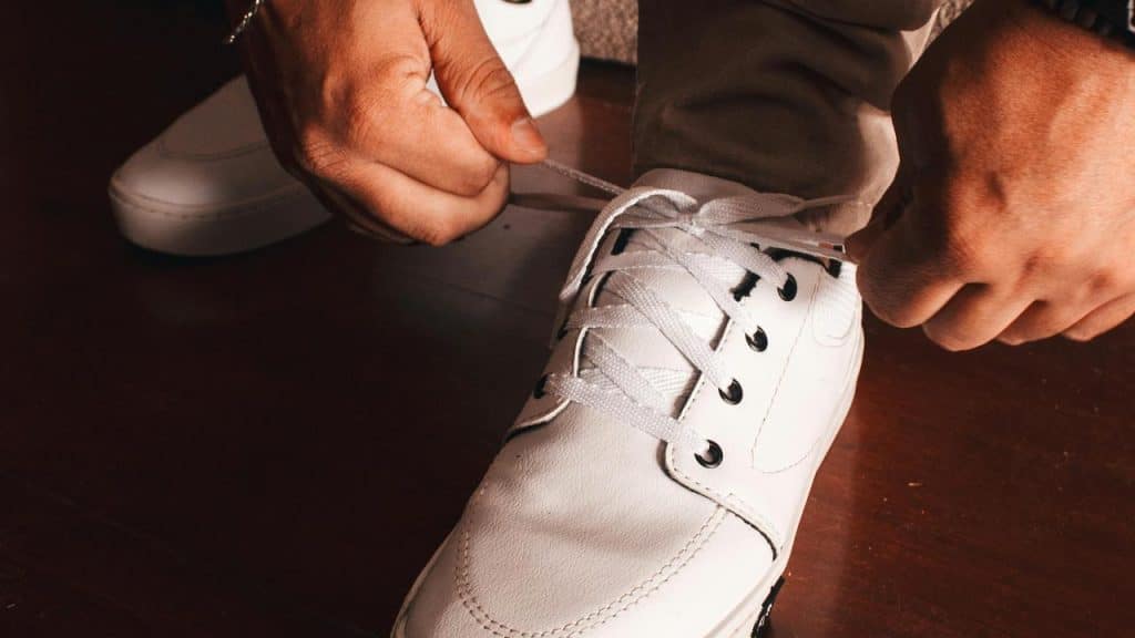 A person tying the laces of white sneakers on a wooden floor.