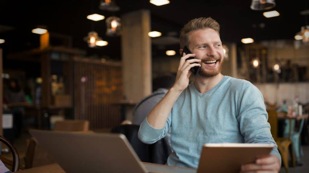 A young happy man having a phone call in a cafe holding a tablet.
