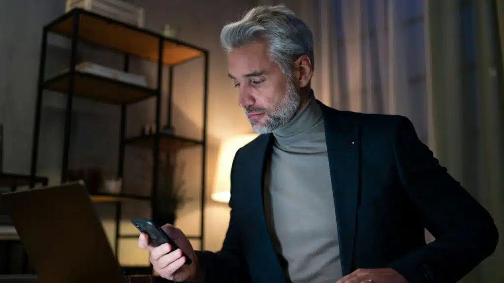 A middle-aged man in a suit jacket and turtleneck looks at his phone in a dimly lit room.