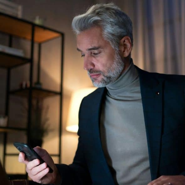 A middle-aged man in a suit jacket and turtleneck looks at his phone in a dimly lit room.