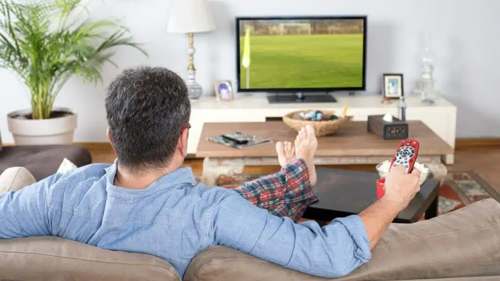 Man relaxing on couch in pajamas, watching TV, holding a remote.