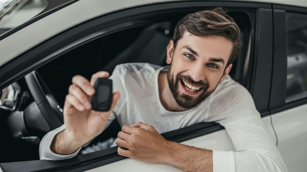 A grinning, bearded man in a white shirt leans out of a car window, holding up a car key fob.