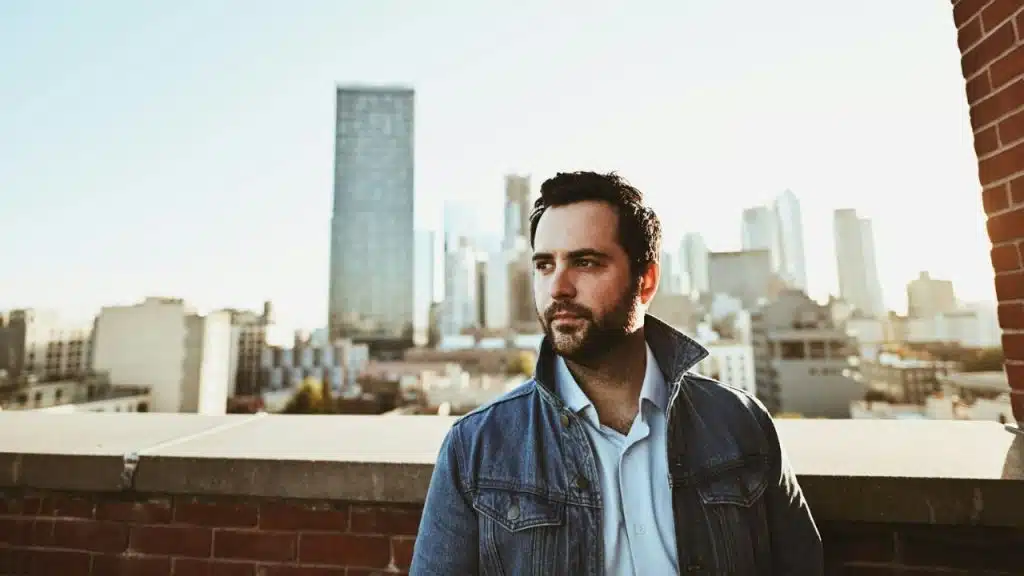 Man in a denim jacket standing on a rooftop with a city skyline behind him.