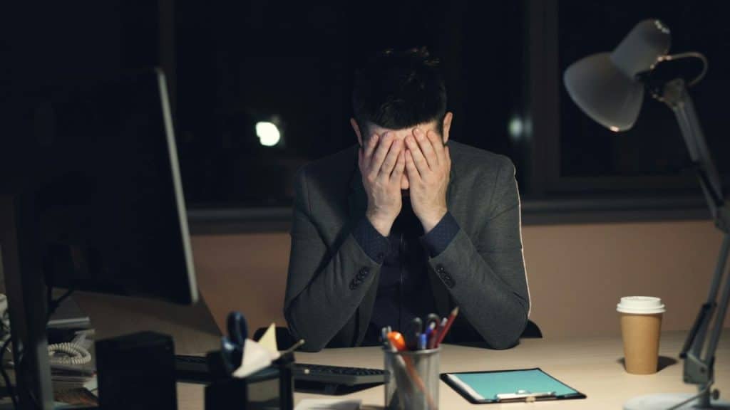 A man in a suit sits at a desk in the dark, covering his face with his hands.