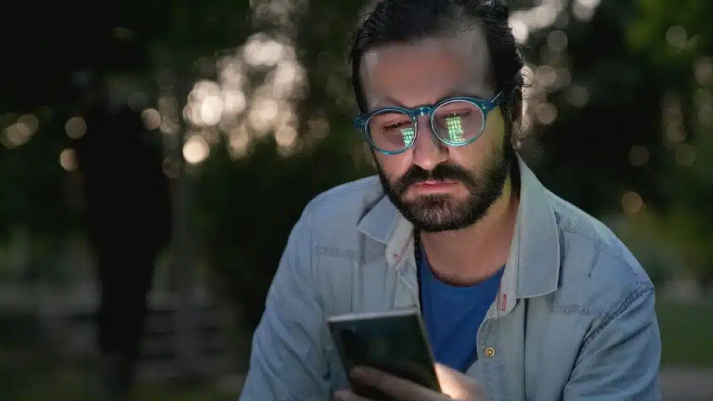 A man with a beard and teal glasses looks down intently at a phone glowing in his hand.