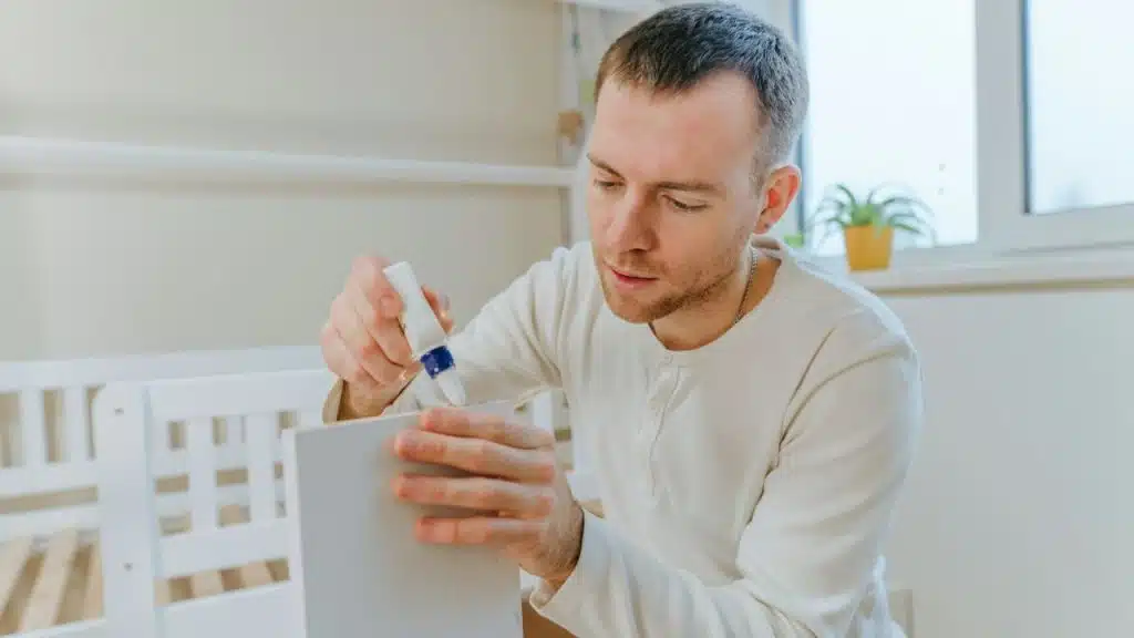A man is gluing a white piece of wood in a bright room with a white crib and large window.