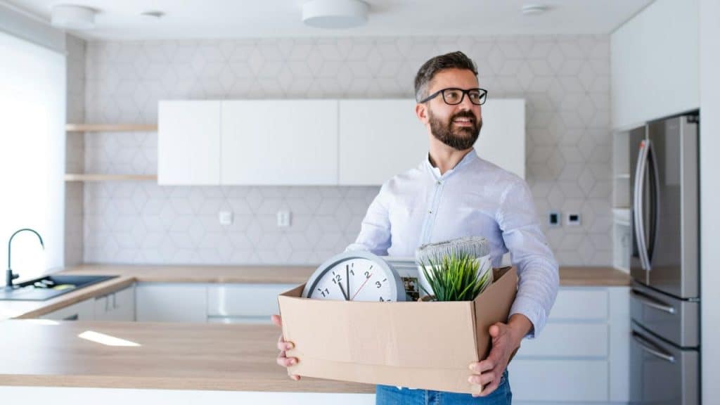 Smiling man with a beard and glasses carries a box of belongings in a modern kitchen.