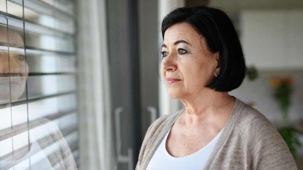 Middle-aged woman with dark hair looks out a window with horizontal blinds.
