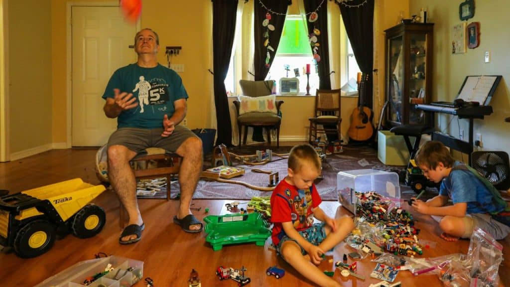 A man sits in a cluttered living room, watching an orange ball he is juggling, while two young boys play with toys on the floor.