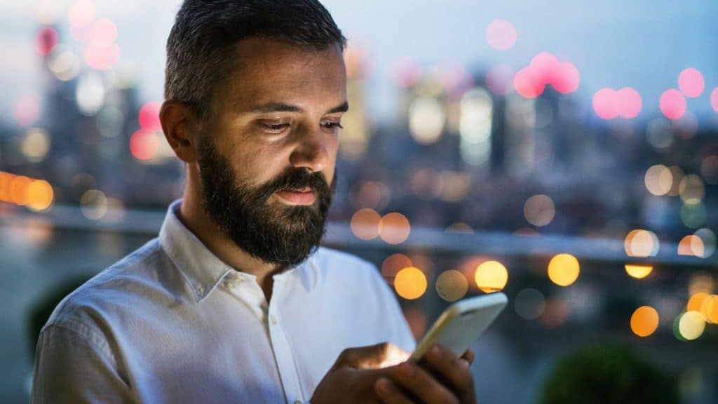 Bearded man in a white shirt looking at his phone with city lights in the background.