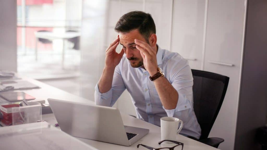 Man with hands on his temples, looking stressed while sitting at a desk with a laptop.