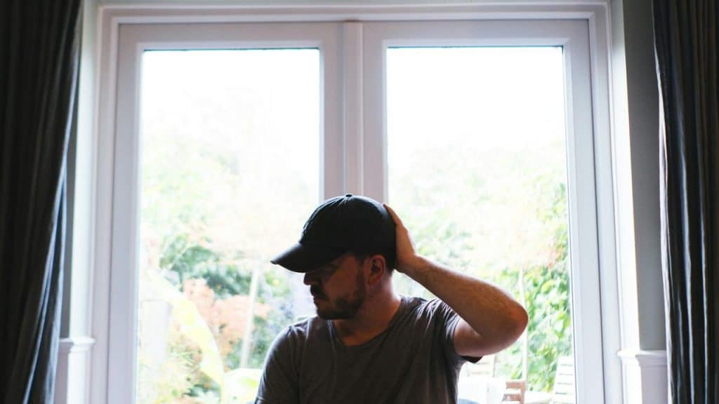 A man wearing a cap sitting indoors with his hand on the back of his head, looking down.