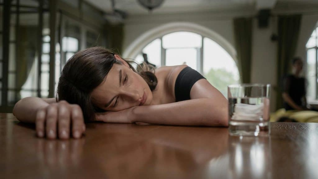 A person resting their head on a table beside a glass of water, appearing tired.