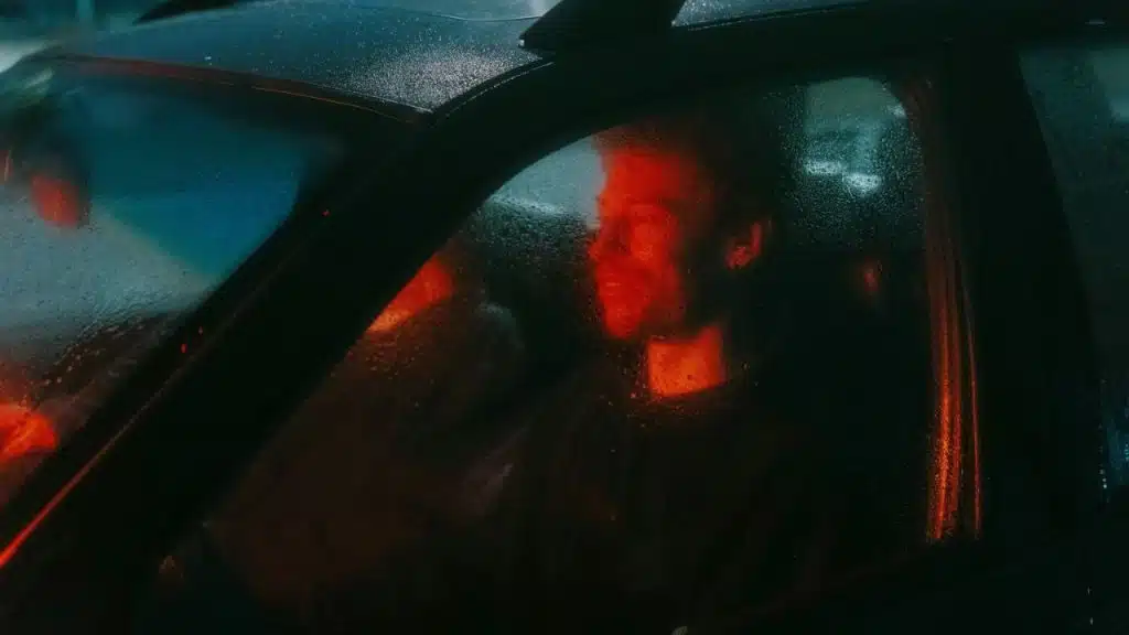 A driver in a car at night, seen through a rainy window with an orange glow on his face.