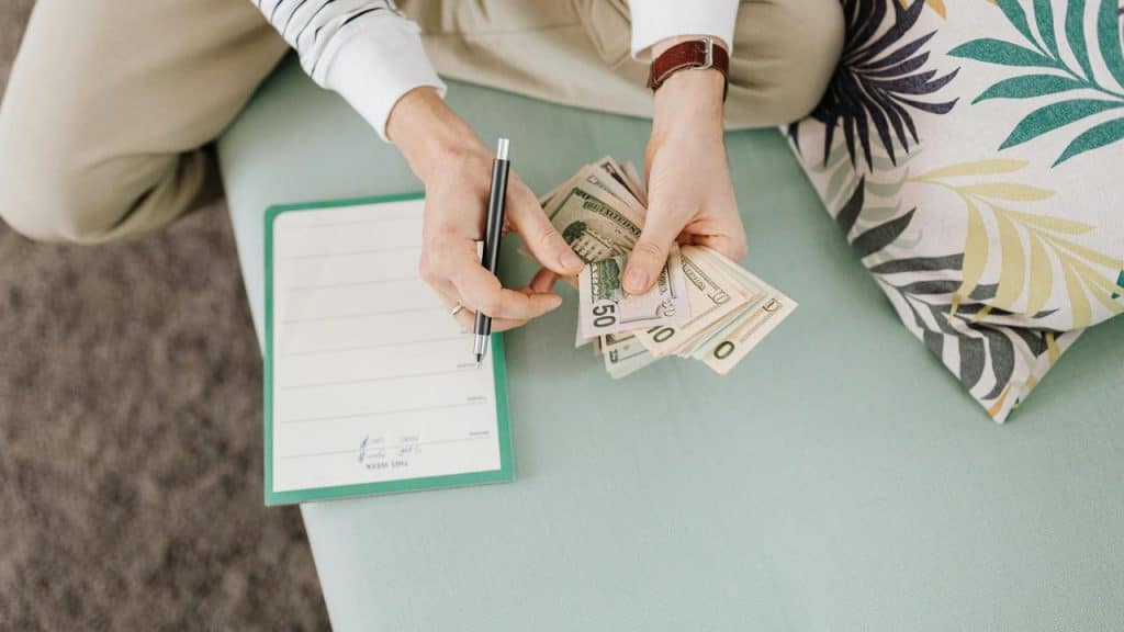 A person holding cash and a pen while sitting next to a notepad.