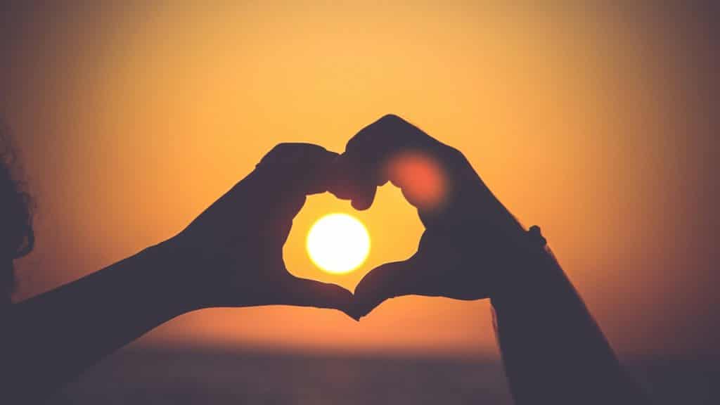 A couple holding up their hands in the form of a heart with the sun framed between. 