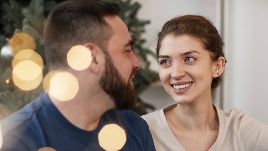A close-up of a happy couple looking at each other, with the woman smiling and bright bokeh lights in the foreground, suggesting a holiday setting