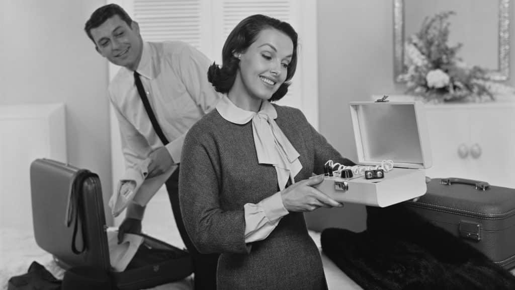 A black and white photo of a smiling woman opening a jewelry box while a man looks on in the background, both standing over suitcases.