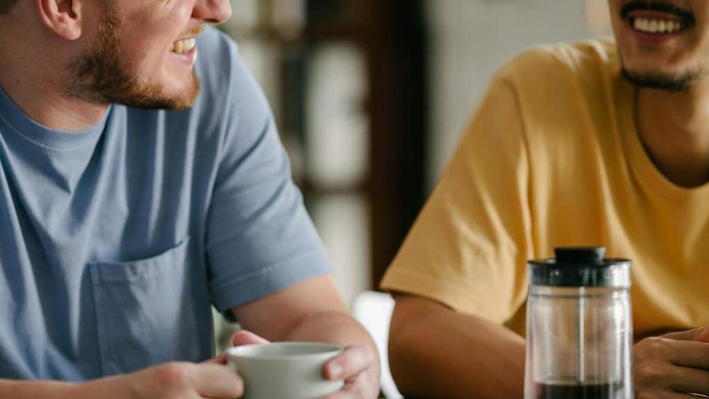 A pair of men sitting at a table smiling and talking over coffee.