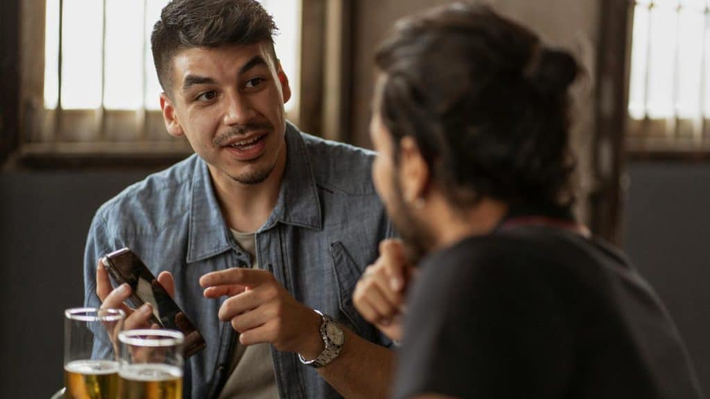 A man holding a phone and talking with a friend over drinks.