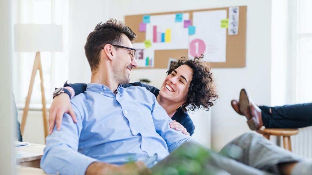 A happy couple embracing and laughing in a bright, modern office, with a corkboard of sticky notes in the background.