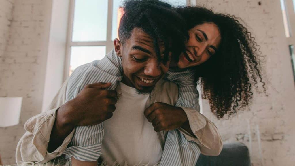 A smiling couple laughing as one gives the other a piggyback ride indoors.