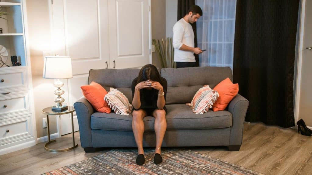 A woman sitting on a couch with her head in her hands while a man stands behind her.