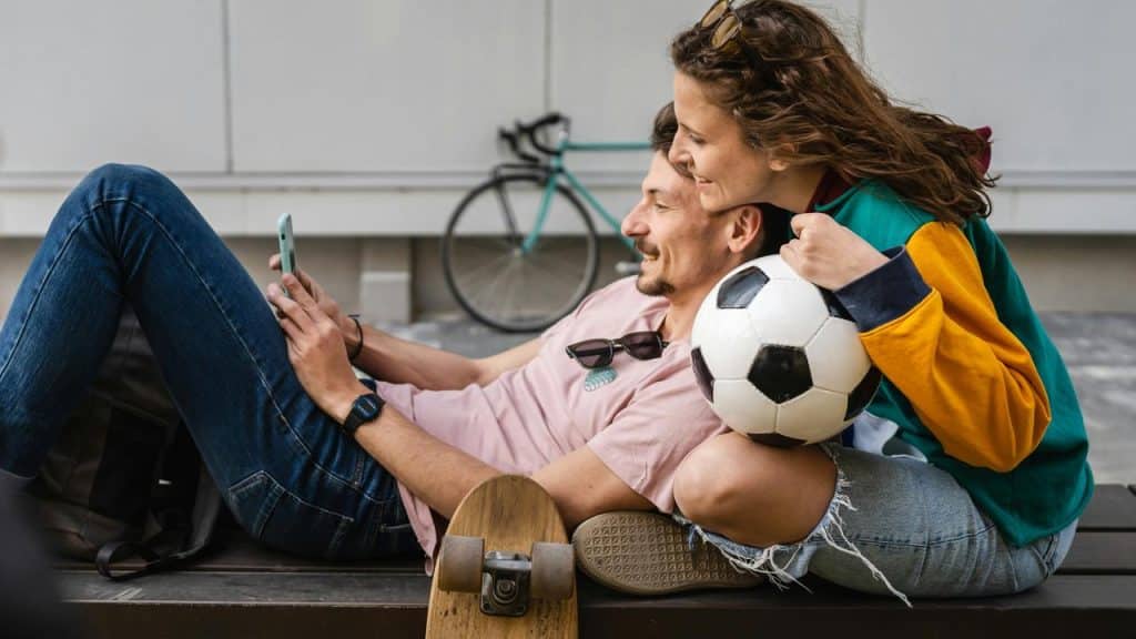 A couple sitting together with a soccer ball and skateboard, smiling at a phone.