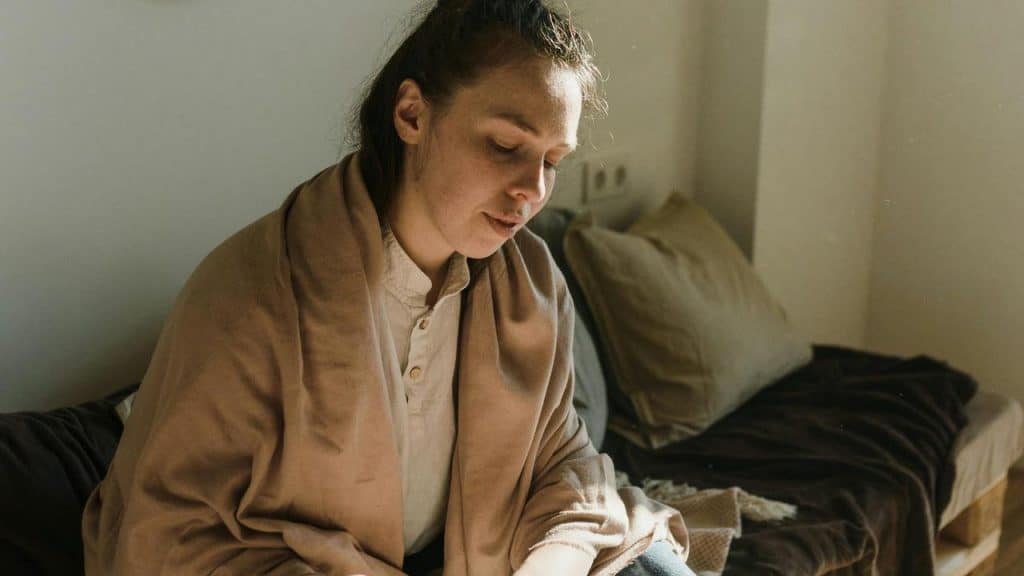 A person sitting on a bed wrapped in a blanket, looking down.