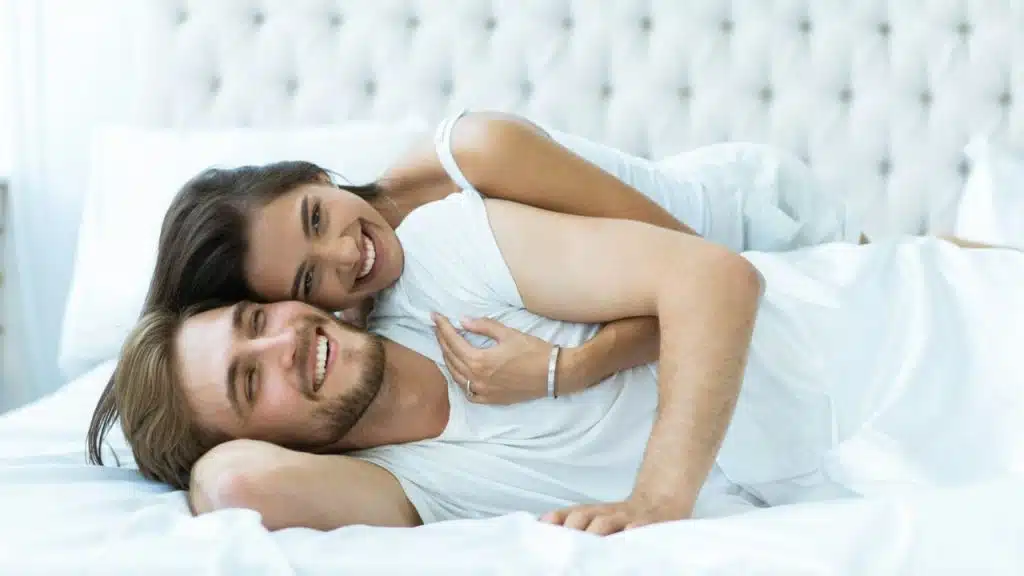 A smiling man is lying on his back while a smiling woman lies on top of him in bed.