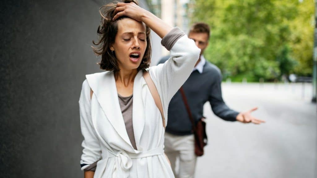 A frustrated woman with her hand on her head walks away from a man who is shrugging and gesturing behind her outdoors.