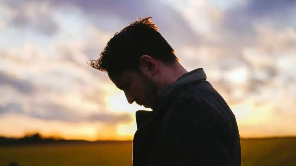 A man is looking down, silhouetted against a setting sun over a field.
