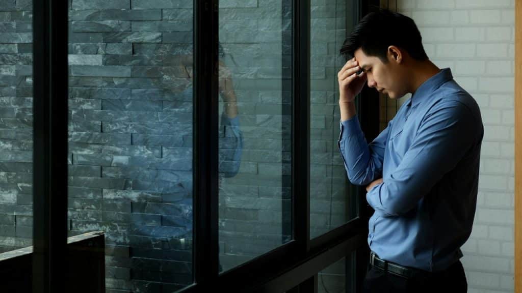 A distressed man in a blue shirt is standing by a window, holding his head.