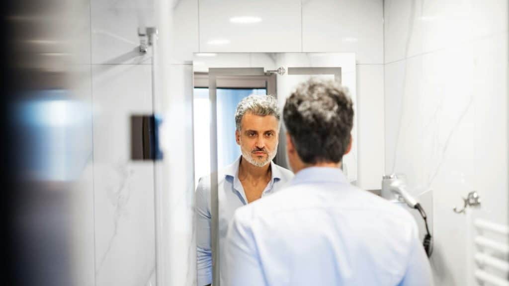 A man with salt and pepper hair is looking at his reflection in a bathroom mirror.