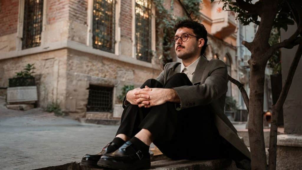 A man with a mustache and glasses sitting outdoors with his hands clasped on his knees.