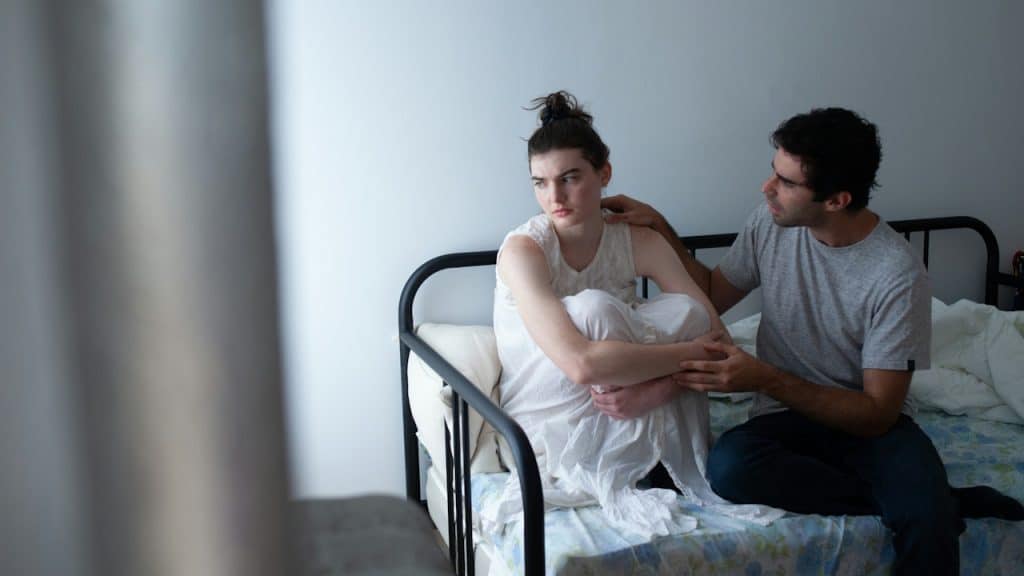 A worried man sits next to a woman curled up and looking away on a bed.